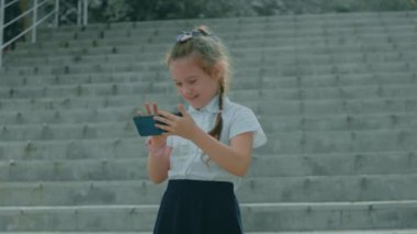 Akıllı telefonla oynayan sevimli bir okul kızı. Neşeli bir 8 yıl. Kız cep telefonuna parmaklarıyla dokunuyor, çizgi film izliyor ve eğitim uygulamalarıyla eğleniyor. Dışarıda parkta bir çocuk