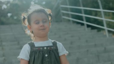 Kafkasya 'nın 2 yıllık güzel portresi. İki örgülü saçı olan küçük bir kız parkta dikiliyor gözleri kapalı kameraya gülümsüyor.