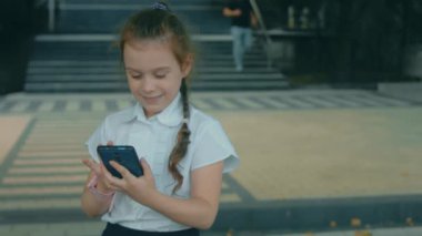 Akıllı telefonla oynayan sevimli bir okul kızı. Neşeli bir 8 yıl. Kız cep telefonuna parmaklarıyla dokunuyor, çizgi film izliyor ve eğitim uygulamalarıyla eğleniyor. Dışarıda parkta bir çocuk