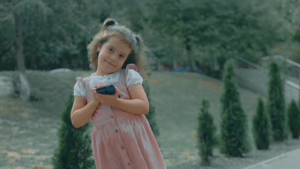 スマートフォンで遊ぶ可愛いプリスクールガールの笑顔 ヘルシー 指で携帯電話を触ったり 漫画を見たり 教育アプリで楽しんだりする少女 屋外で公園にいる子供たち — ストック動画
