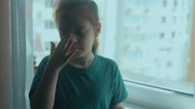 Pencerenin kenarında tek başına oturmuş ağlayan ve gözyaşlarını silen üzgün küçük kız. Yalnız, endişeli çocuk pencerede oturuyor.