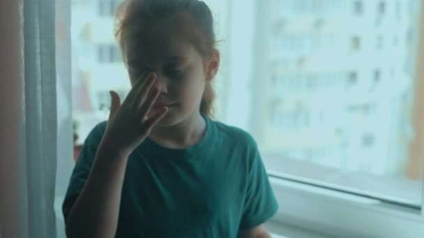 悲しい少女が泣いて涙を拭きながら窓に一人で座っている 窓に座っている孤独な心配の子供 — ストック動画
