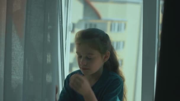 伤心的小女孩独自坐在窗台上哭泣着 用手擦拭着眼泪 一个孤独的孩子坐在窗边 — 图库视频影像
