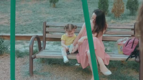 妈妈和女儿坐在操场上的长椅上 白人年轻快乐的女人用小石头擦拭她的小女孩的鞋子 — 图库视频影像