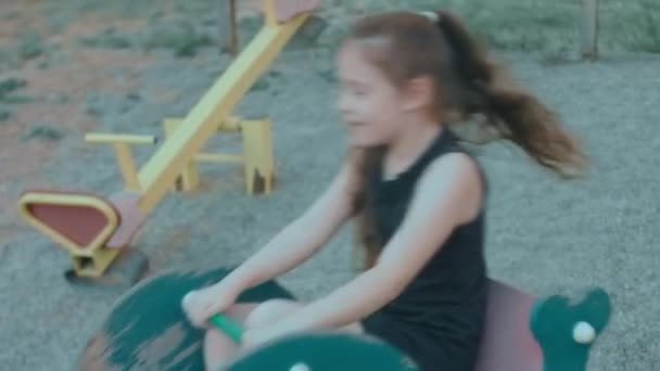 ロッキング馬の遊び場で楽しんでいる美しい学校の女の子 晴れた夏の日にアウトドアを楽しむ — ストック動画