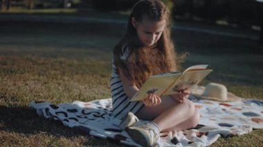 Battaniyeye oturmuş, doğadaki piknikte kitap okuyan tatlı kıvırcık kız. Yaz, tatil, boş zaman ve mutlu çocukluk konsepti.
