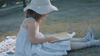 Güneş şapkası takan şirin kıvırcık kız battaniyeye oturmuş ve doğadaki bir piknikte kitap okuyor. Yaz, tatil, boş zaman ve mutlu çocukluk konsepti.