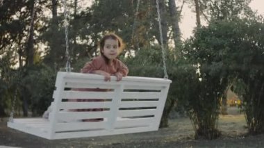 Beyaz bir salıncakta, ağaçlarla çevrili arka planda küçük bir kız eğleniyor. Anaokulu öğrencisi bahçede salıncakta sallanıyor.