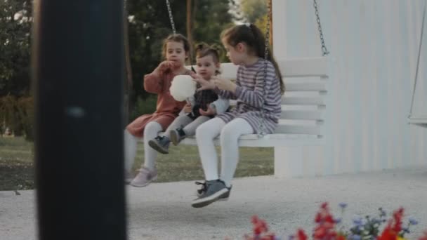 三个小女孩在一个安静的 绿树成荫的公园里玩着一个白色的秋千 姐姐们在后院的长椅秋千上吃棉花糖 — 图库视频影像
