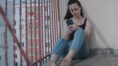 Şehir manzarası güzel olan balkonda otururken akıllı telefona bakan bir kadının yakın çekimi. Kadın video izliyor, sosyal ağda sohbet ediyor..