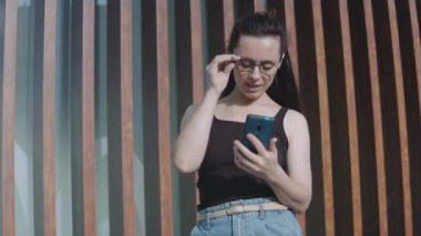 Gözlüklü genç bir kadın modern bir binanın yanında tek başına duruyor, gülümsüyor ve cep telefonu kullanıyor, ekrana bakıyor, birileriyle konuşuyor, uygulama sohbetinde mesajlar gönderiyor.
