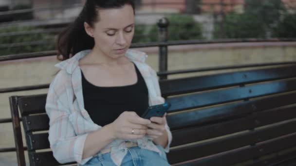 ベンチに一人で座り 携帯電話を使い 画面を見たり テキストを送ったり アプリチャットでメッセージを送ったりする若い女性 — ストック動画