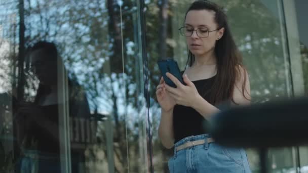 建物の前に一人で立っている眼鏡を持つ若い女性 携帯電話を使用して 画面を見て テキスト アプリチャットでメッセージを送信 — ストック動画