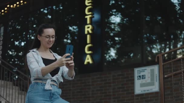 建物の前に一人で立っている眼鏡を持つ若い女性 携帯電話を使用して 画面を見る セルフィーを作る テキスト アプリチャットでメッセージを送信 — ストック動画