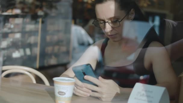 接近拿着手机的女人 在喝咖啡休息的时候看手机上的视频 从外面看 — 图库视频影像