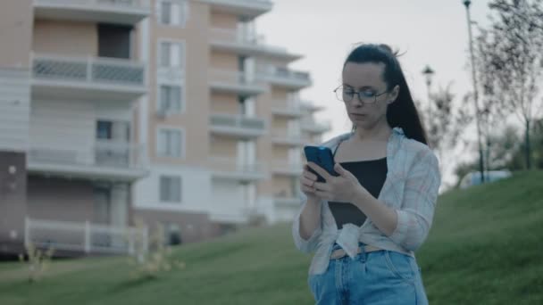 住宅地に一人で立っている眼鏡を持つ若い女性 携帯電話を使用して 画面を見て テキスト アプリチャットでメッセージを送信 — ストック動画