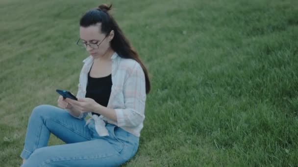 グリーン芝の上に座っている眼鏡を持つ若い女性 携帯電話を使用して 画面を見て テキスト アプリチャットでメッセージを送信 — ストック動画