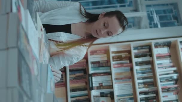 バーティカルシューティング 笑顔の若い女性は本屋で彼女の自由な時間を過ごしています 買うために新しい本を探しています — ストック動画
