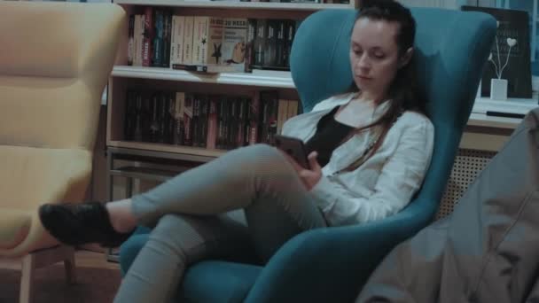年轻的女人正在书店度过她的闲暇时光 坐在扶手椅上放松 还在看智能手机 — 图库视频影像