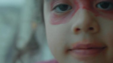 Neşeli, kendine güvenen küçük beyaz kız yüzünü ruj ve göz farı ile boyamış. Küçük kız sırf eğlence olsun diye dudaklarını ve göz kapaklarını maske olarak boyadı.