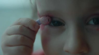 Neşeli, kendine güvenen beyaz çocuk yüzünü ruj ve göz farı ile boyamış. Küçük kız eğlence olsun diye dudaklarını ve göz kapaklarını boyuyor. Kapat..