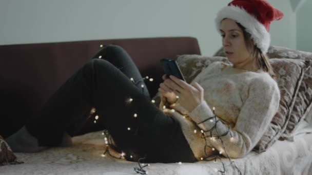 スマートフォンを持っている赤いサンタの帽子の女性 自宅でクリスマスを祝う若い女性 携帯電話を使用して携帯電話を待っています — ストック動画
