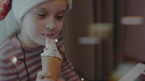 クリスマスパーティーでアイスクリームを食べるサンタの帽子をかぶっているかわいい少女 周りのクリスマスライト — ストック動画