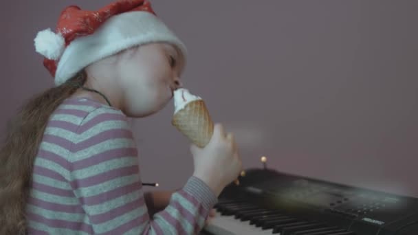 クリスマスパーティーでアイスクリームを食べるサンタの帽子をかぶっているかわいい少女 ピアノを弾き 楽しんでいる 周りのクリスマスライト — ストック動画