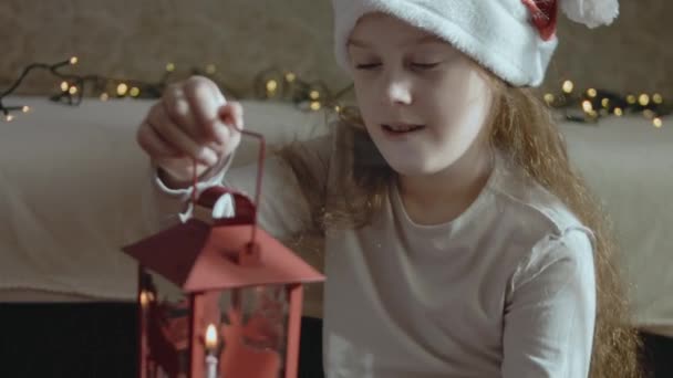 サンタスの帽子をかぶったかわいい少女が ろうそくを燃やしたランタンを抱えています クリスマスのコンセプト — ストック動画