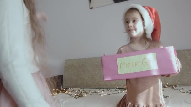 謎のギフトボックスを持っている幸せな少女 別の女の子は 謎の箱の中にあるものを推測するジェスチャーを示しています クリスマスイブで遊ぶ小さな女の子 — ストック動画