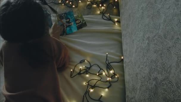 一个小女孩在圣诞夜看着一张旧照片 圣诞灯亮着圣诞节和休闲的概念 后视镜 — 图库视频影像