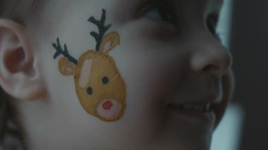 Yüzünde Noel temalı resim olan neşeli küçük kız. Yüzünde kar taneleri ve ren geyiği resmi var. Noel konsepti