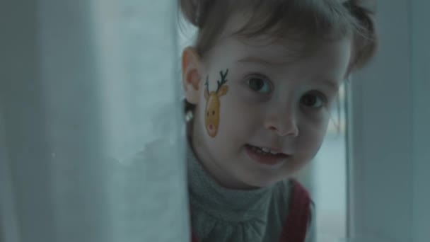 彼女の顔にクリスマスをテーマにした絵を描いた陽気な幼児の少女 カーテンの後ろに隠れている小さな女の子がピカブーを演奏します クリスマスコンセプト — ストック動画