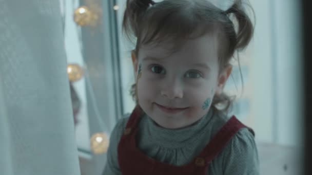 彼女の顔にクリスマスをテーマにした絵を描いた陽気な幼児の少女 スノーフレークとトナカイの絵が顔に描かれています クリスマスコンセプト スローモーション — ストック動画