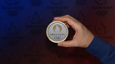 Bir erkek el, üzerinde mavi-kırmızı gradyan arka planda Paris 2024 Olimpiyat Oyunları logosu bulunan yuvarlak altın çerçeveli bir plaket tutuyor..