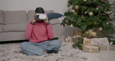 Yeni yıl hediyesi. Sanal gerçeklik gözlüğü takan genç bir kadın ilk kez Noel ağacının yanındaki rahat koltukta oturuyor. Mutlu bir kadın Noel tatilinde 3D VR gözlüklerini test ediyor..