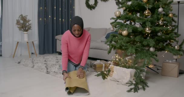 ギフトラッピング 黒人の若い女性はクリスマスプレゼントを家の木の下に置く準備ができている — ストック動画