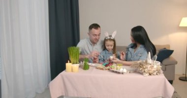 Tavşan kulaklı şirin küçük kız ailesiyle vakit geçiriyor. Annem ve babam paskalya yumurtası boyuyor, boya fırçalarıyla oynuyorlar..