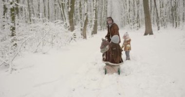 Noel tatilleri, kış orman düzenlemeleri. İki çocuklu aile kızak çekiyor ve kış ormanlarında karlı patika boyunca yürüyorlar.