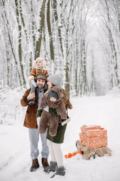 stock image Parents with their children having fun in forest on snow day. Happy toddler daughter and baby son wearing warm clothes. Gift boxes standing next to them.