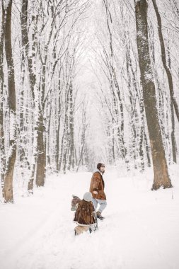 Anne-babalar ve küçük oğlu kar tatilinde ormanda eğleniyorlar. Küçük kızımız ve oğlumuz sıcak kıyafetler giydiği için çok mutluyuz. Adam karısını ve oğlunu kızakla gezdiriyor..