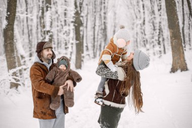 Aileler çocuklarıyla birlikte kar tatilinde ormanda eğleniyorlar. Küçük kızımız ve oğlumuz sıcak kıyafetler giydiği için çok mutluyuz. Anne ve baba çocuklarını ellerinde tutuyorlar..