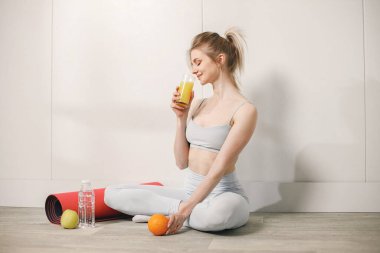 Bir bardak meyve suyuyla yerde oturan ve poz veren genç bir kadın. Gri bluzlu ve tozlu bir kadın. Kırmızı hasırın yanında oturan kadın, bir şişe su, bir portakal ve bir elma..