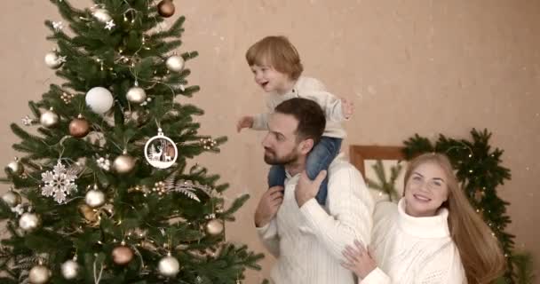 自宅でクリスマスツリーを飾る彼の小さな息子と妻と若い男 お父さんがリビングルームで新年の木を準備するのを助けるCurios魅力的な息子 — ストック動画