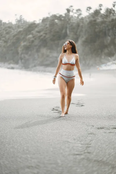 黒砂のビーチを歩くブルネットの女の子は バリでかわいい水着を着てインドネシア — ストック写真