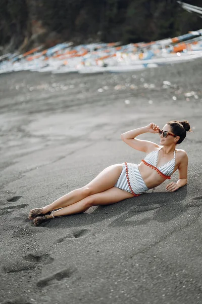 ブルネットの女の子はバリ島でかわいい水着を着て隠された黒い砂浜に横たわっていますインドネシア — ストック写真