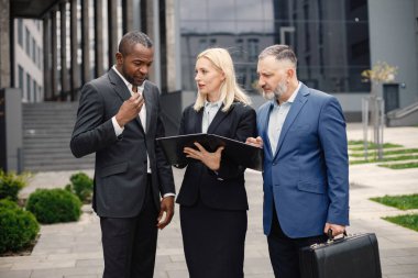 Modern ofis binasının önünde kendinden emin iş adamları konuşuyor. Belgelere bakan siyah ve beyaz erkekler ve sarışın kadınlar. Bankacılık ve finans piyasası kavramı.