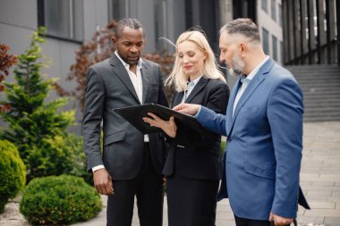 Modern ofis binasının önünde kendinden emin iş adamları konuşuyor. Belgelere bakan siyah ve beyaz erkekler ve sarışın kadınlar. Bankacılık ve finans piyasası kavramı.
