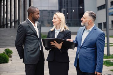 Modern ofis binasının önünde kendinden emin iş adamları konuşuyor. Belgelere bakan siyah ve beyaz erkekler ve sarışın kadınlar. Bankacılık ve finans piyasası kavramı.