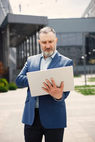Caucasian man wearing blue jacket and using a laptop. Confident businessman standing in front of modern office building. Banking and financial market concept.